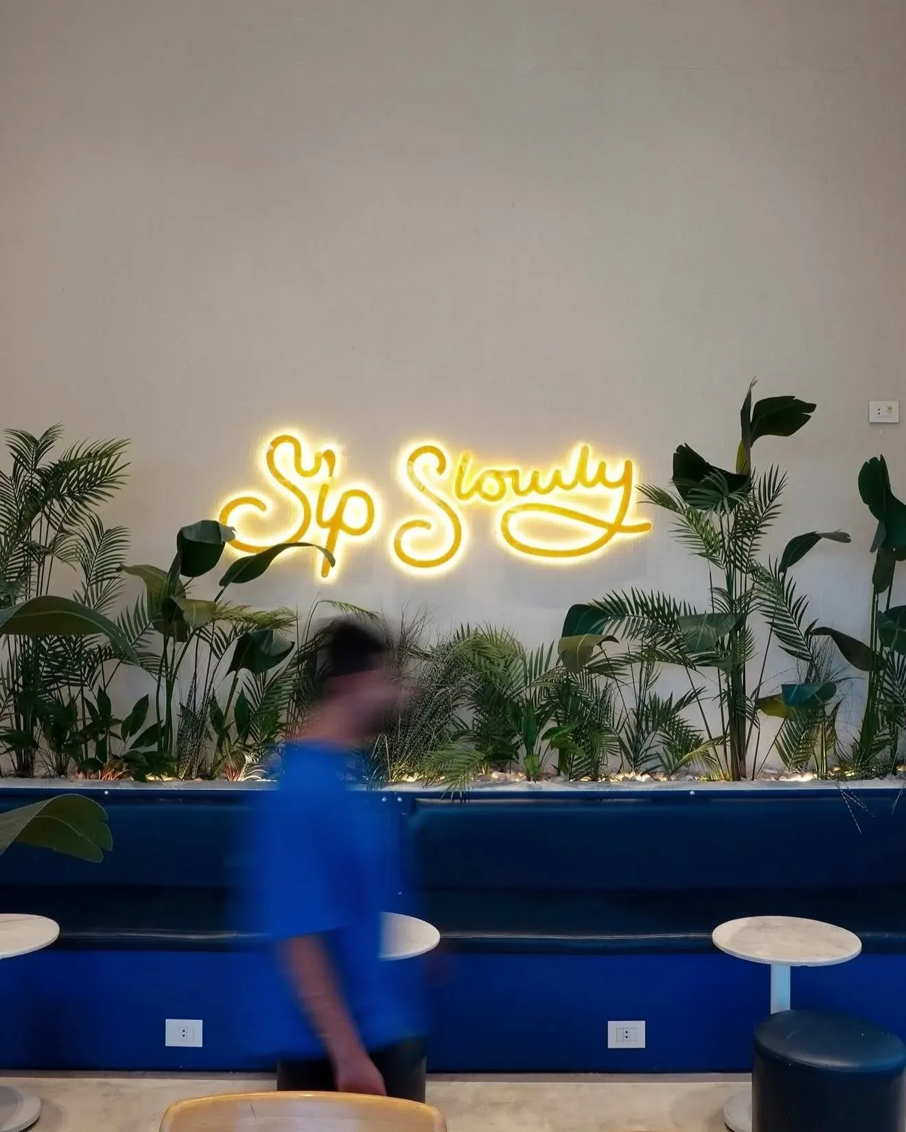 Yellow neon Sip Slowly sign above a blue banquette and plants