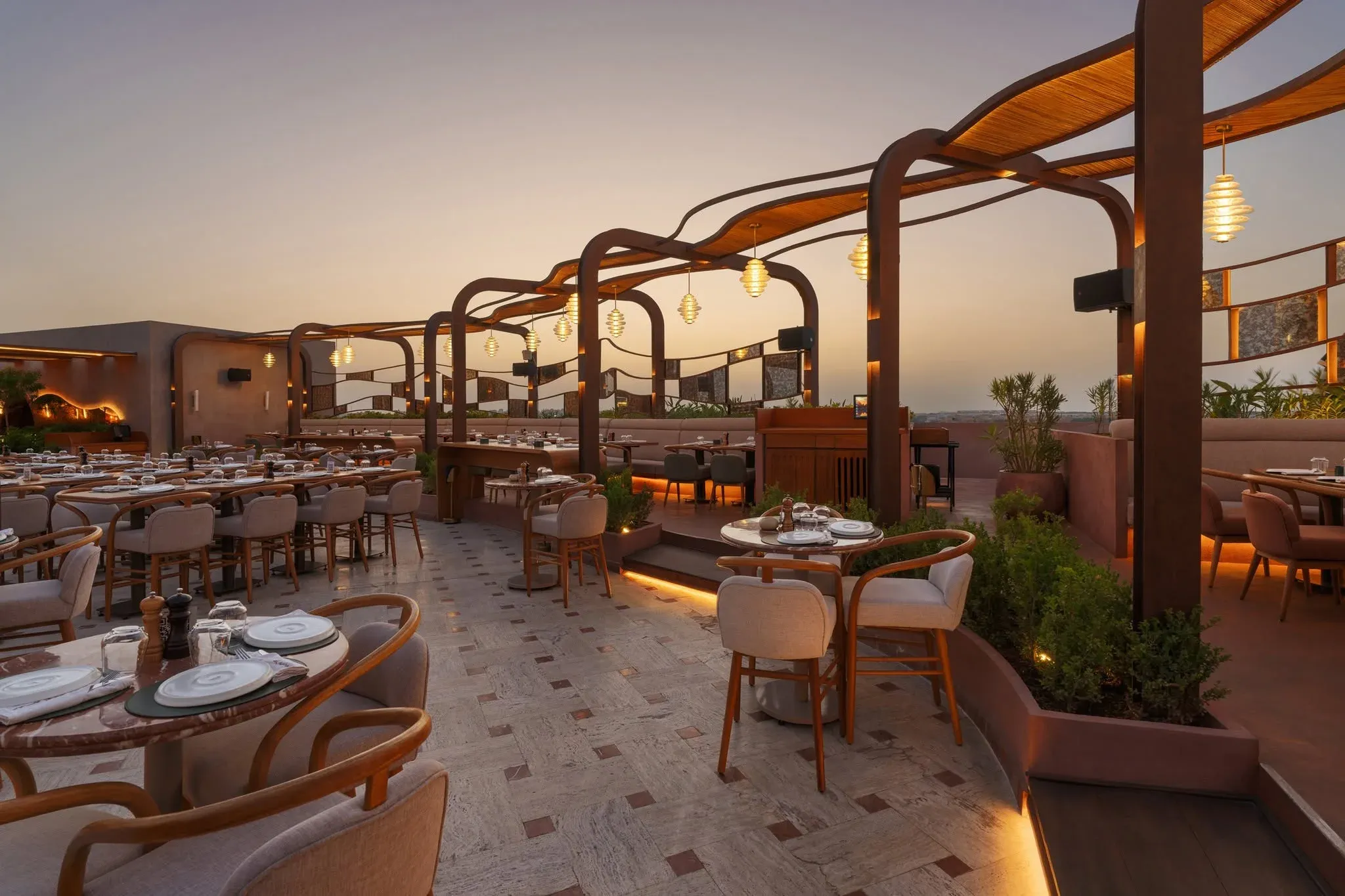 Dining tables and chairs under steel arches and pendant lights at dusk